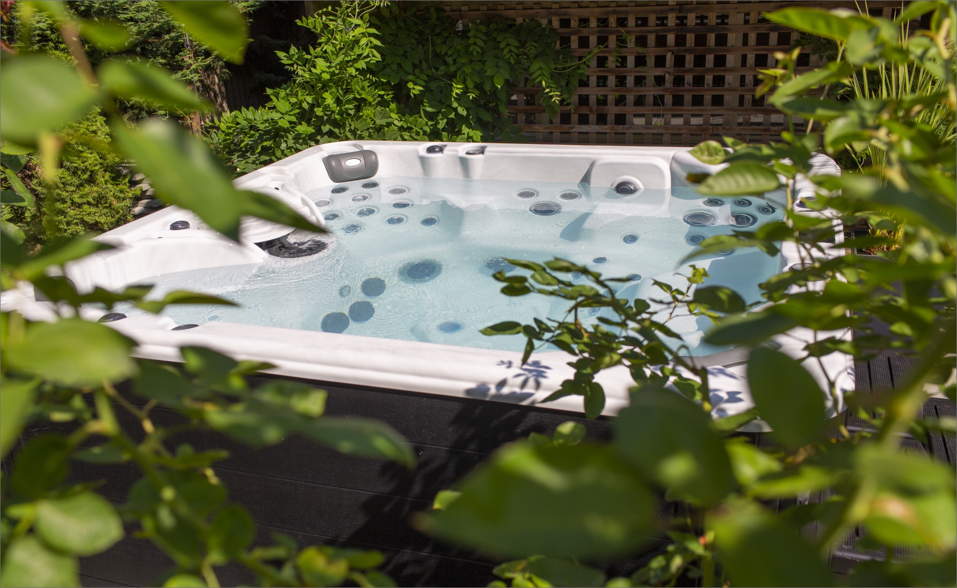 A white hot tub filled with clear water is surrounded by lush green plants and foliage. The hot tub has multiple water jets and is situated in an outdoor garden area, partially enclosed by a wooden lattice fence in the beautiful setting of Red Deer, Alberta.