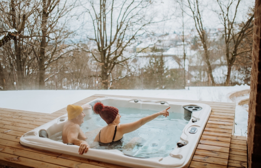 Two people relax in an outdoor hot tub on a wooden deck overlooking a snowy landscape. They are wearing winter hats, and one is pointing toward the distance, possibly spotting a Red Deer. The scene is framed by bare trees and snow-covered ground, creating a cozy and scenic winter atmosphere.