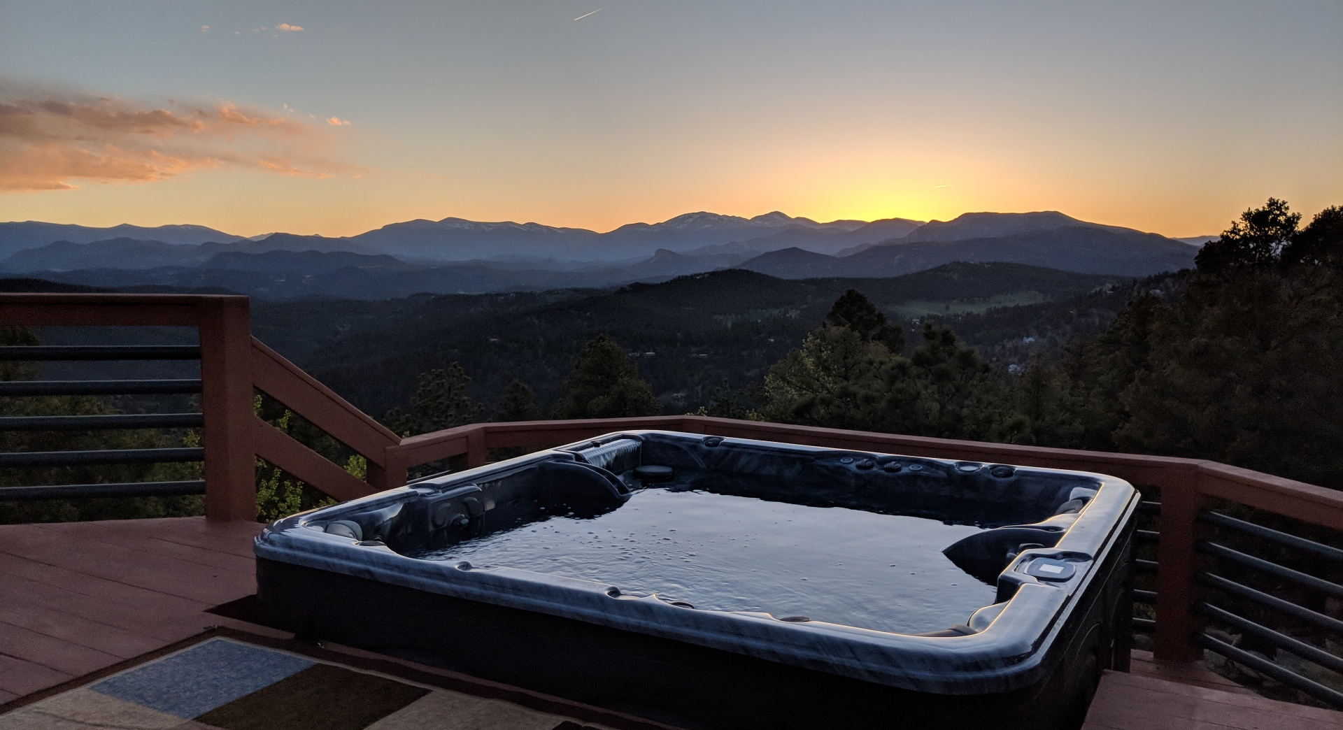 A serene sunset view of a hot tub on a wooden deck overlooking the vast mountain ranges of Central Alberta, Canada. The sky is a blend of warm oranges, yellows, and dusky blues, casting a peaceful glow over the landscape. The hot tub's calm water reflects the colors of the evening sky.